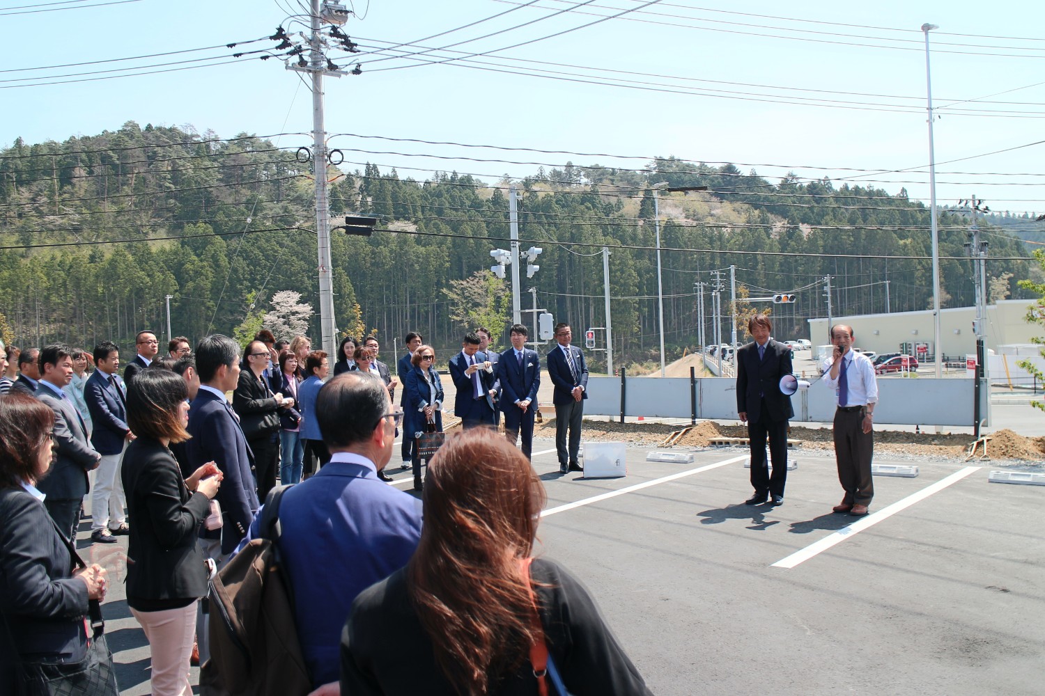 スーツ姿の人々が屋外の舗装された駐車場に集まり、前方で2人の男性が拡声器を使って話している様子を周囲の人たちが真剣な表情で見守っている開会式のような場面の写真