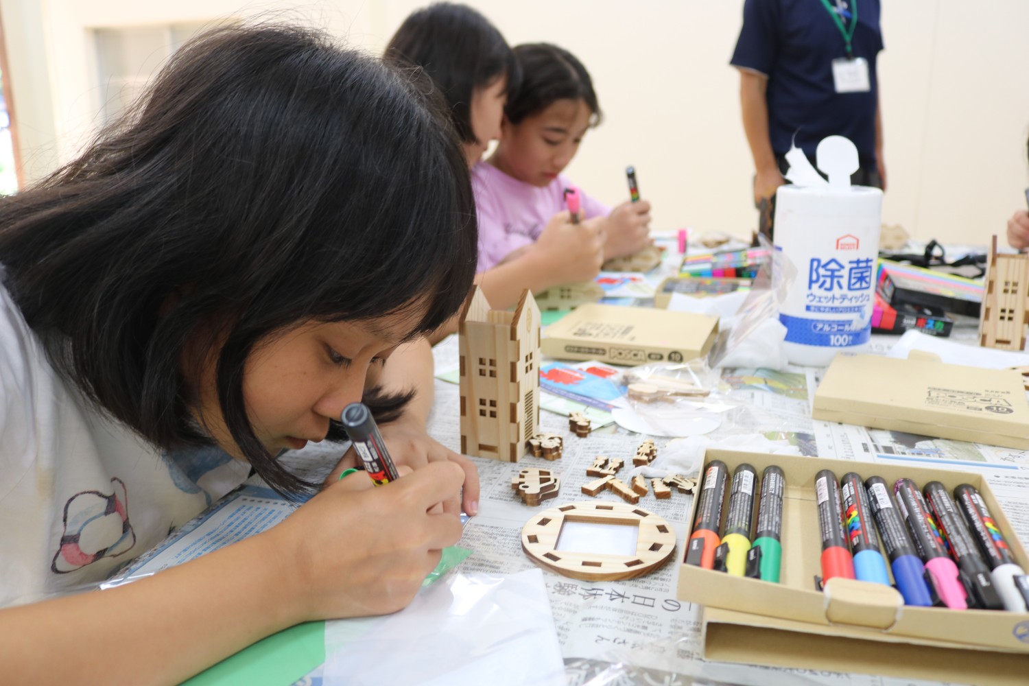 数人の子どもたちが机の上で木製の工作キットを使って作業しており、手前の女の子が集中して紙に色を塗っている写真