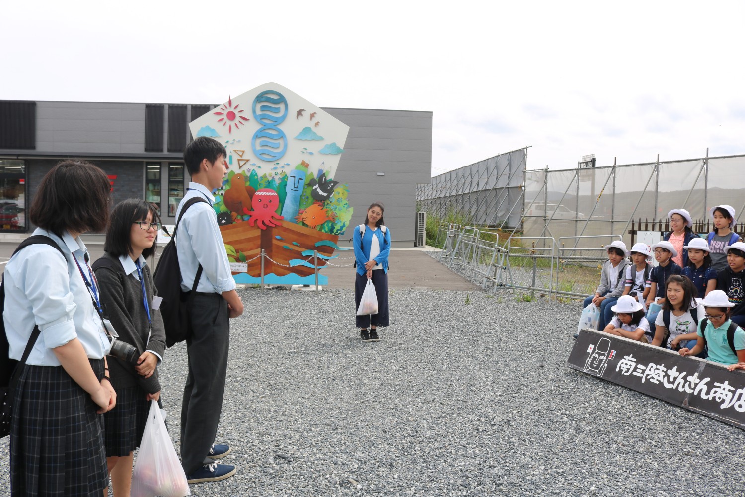 南三陸さんさん商店街のカラフルな壁画の前で、左側に立つ制服姿の志津川高校生と右側に座る白い帽子をかぶった小学生たちが向き合いながら交流している様子が写っている写真