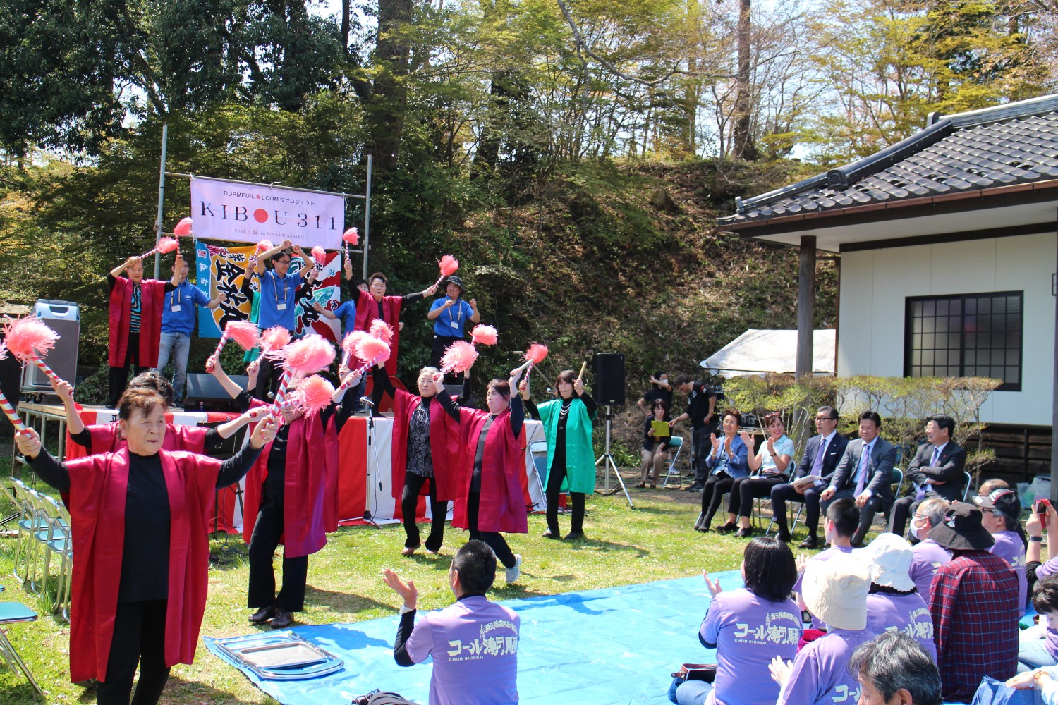 椅子に座っている来賓者や青いビニールシートが敷かれた客席では多くの人が座って拍手を送っている屋外ステージの前で、赤や緑の衣装を着た高齢の女性たちが両手にピンクのポンポンを持って踊っており、後方には青いポロシャツの大学生たちが一緒に踊っているステージ発表の写真