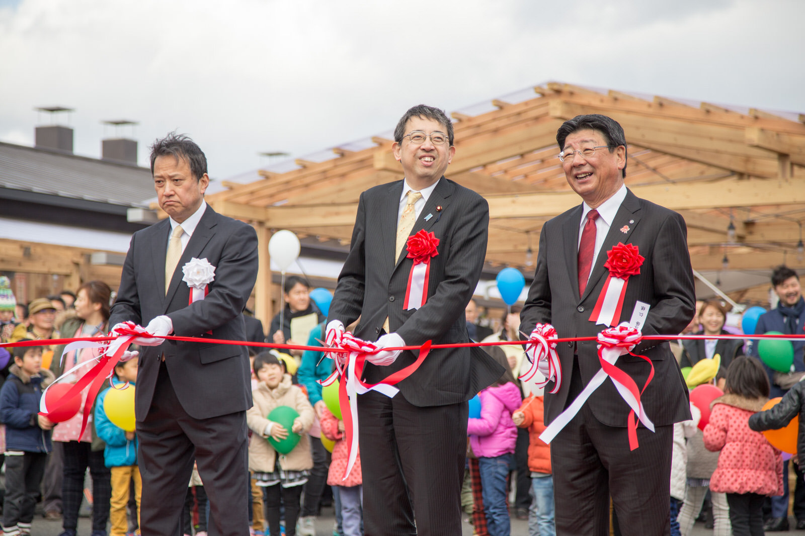 後ろには子どもたちが風船を持って並んでいるなか、赤いリボンを持ったスーツ姿の3人の関係者の男性がテープカットをしている様子の写真
