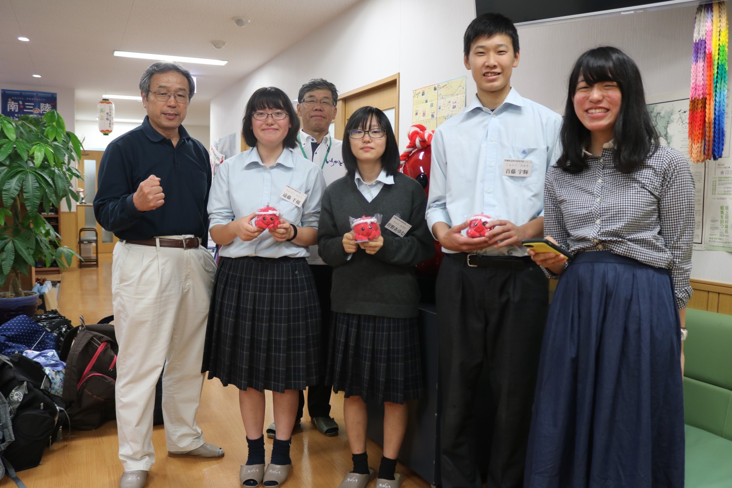 千羽鶴が飾られた建物の廊下で、手には赤いオクトパス君のぬいぐるみを持っている学生服を着た男女と、2人の男性が並んで記念撮影をしている写真