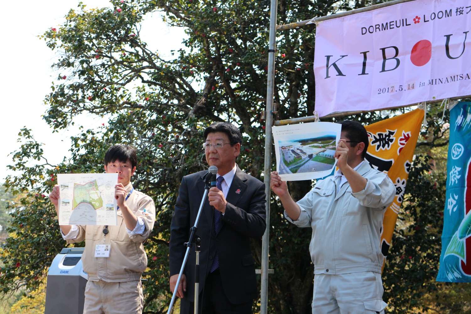 背後にはピンク色の横断幕が掲げられている屋外のステージ上で、作業服を着た2人が地図や完成予想図のパネルを掲げている中央でスーツ姿の佐藤町長がマイクを手に説明している様子の写真