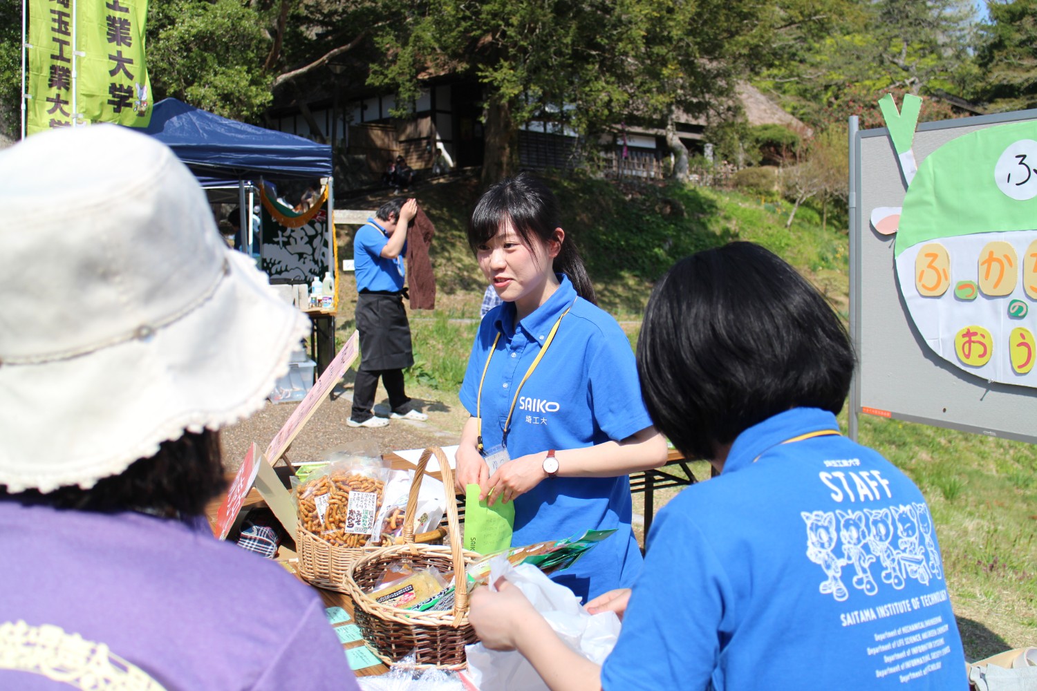 丘の上に茅葺屋根の建物が建っている前の広場で、青いポロシャツを着た女子大生がテーブルに並べられたお菓子の入ったカゴを前にして来場者と話をしている写真