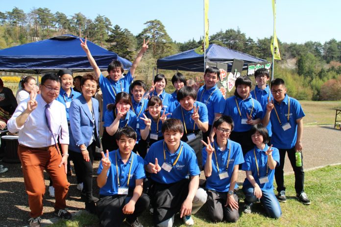 周囲を木に囲まれた青色のテントが設置された晴れた日の屋外で、白いシャツを着た男性と水色のジャケットを着た女性、お揃いの青いポロシャツを着た若者たち16人がカメラに向かって笑顔でピースサインをしている集合写真