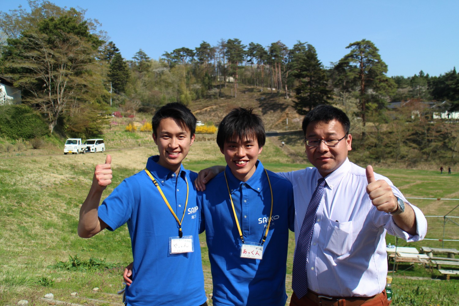 木々が立つ小さな丘と草原の中で、青いポロシャツを着た2人の大学生とシャツとネクタイ姿の准教授が肩を組み親指を立てて笑顔を見せている記念撮影の写真