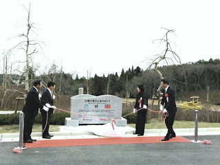 葉の落ちた木々と針葉樹が混在する森を背景に、正装した4人の人物が石碑の周りに立ち、石碑の除幕式を行っている様子を写した式典の写真