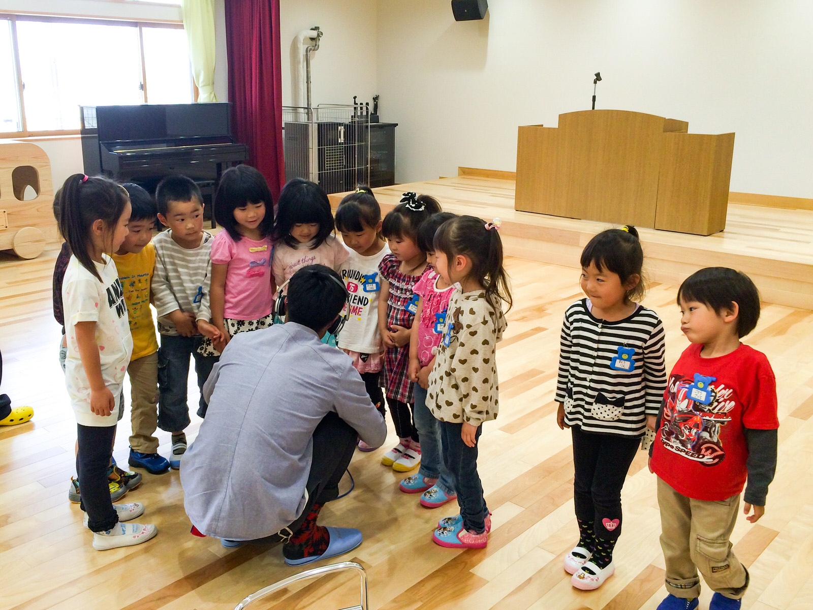 演台が置かれたステージがある部屋で、しゃがんでいるヘッドホンをつけた男性の周りに11人の園児たちが興味津々に集まっている様子の写真