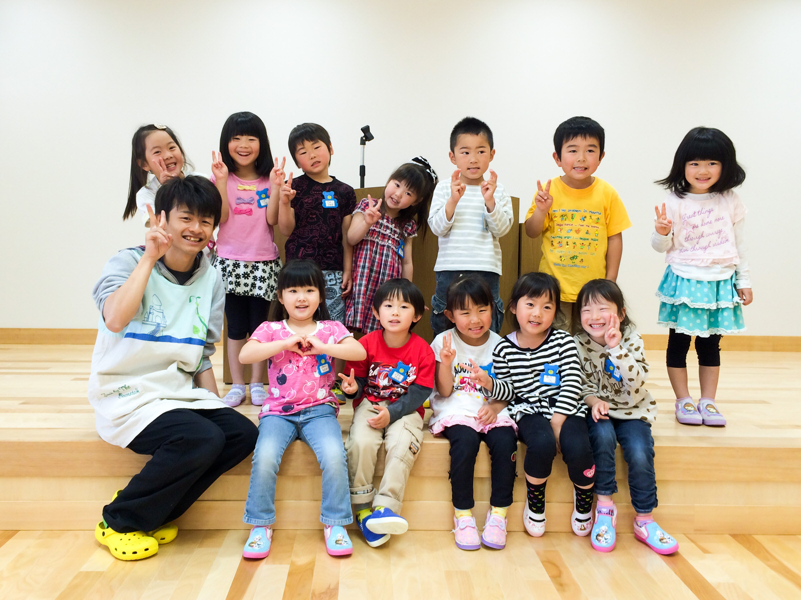 ステージの端に保育士の男性と5人の園児が座り、その後ろに7人の園児が立ってピースサインなどのポーズをとって写っている集合写真