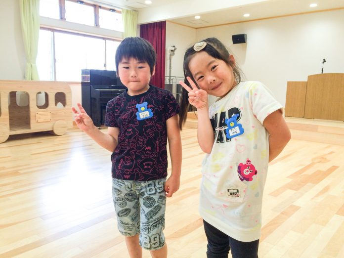 演台が置かれたステージがある部屋で、男女の園児2人が横に並んで立ち、ピースサインをしている写真
