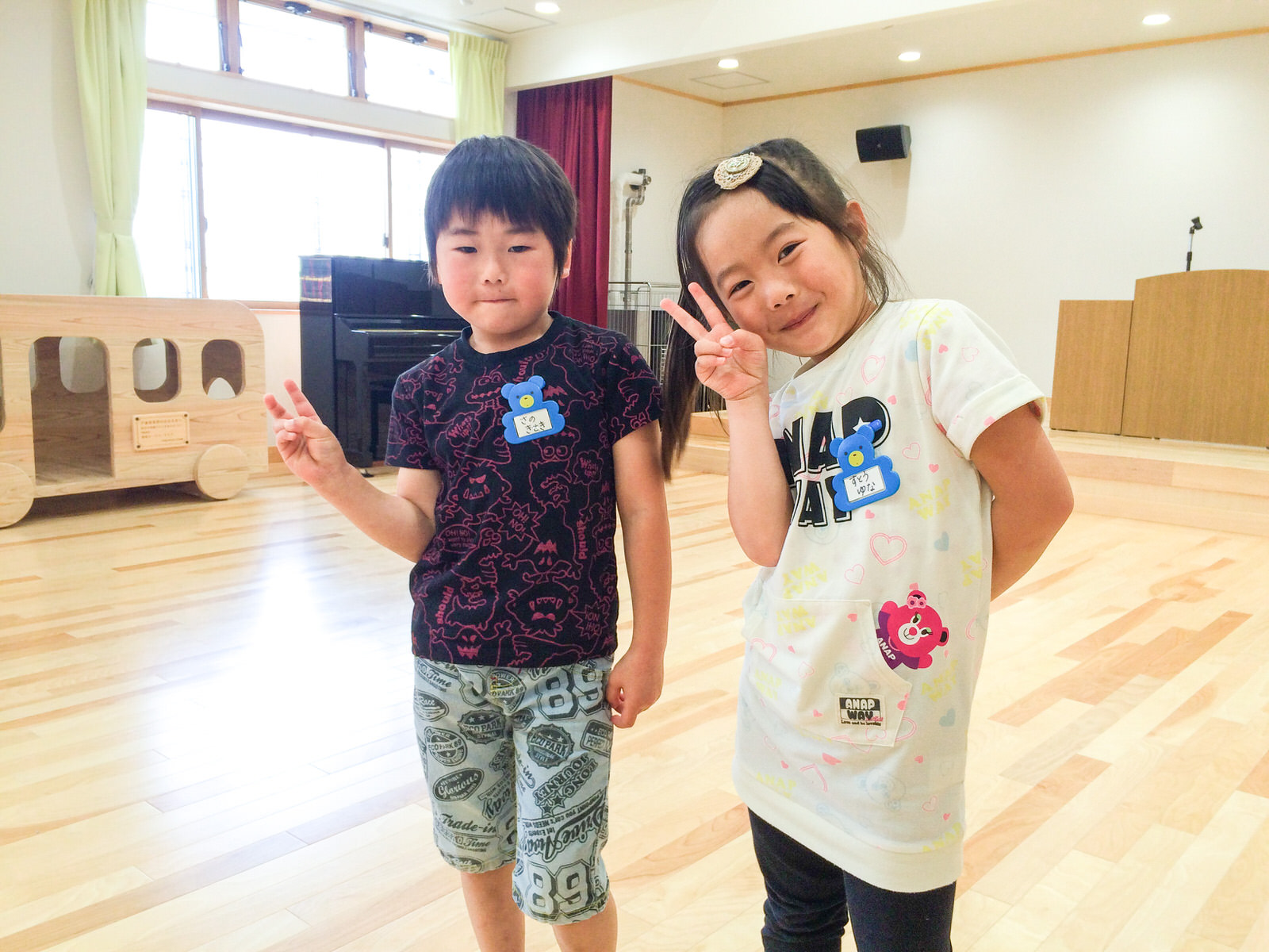 演台が置かれたステージがある部屋で、男女の園児2人が横に並んで立ち、ピースサインをしている写真