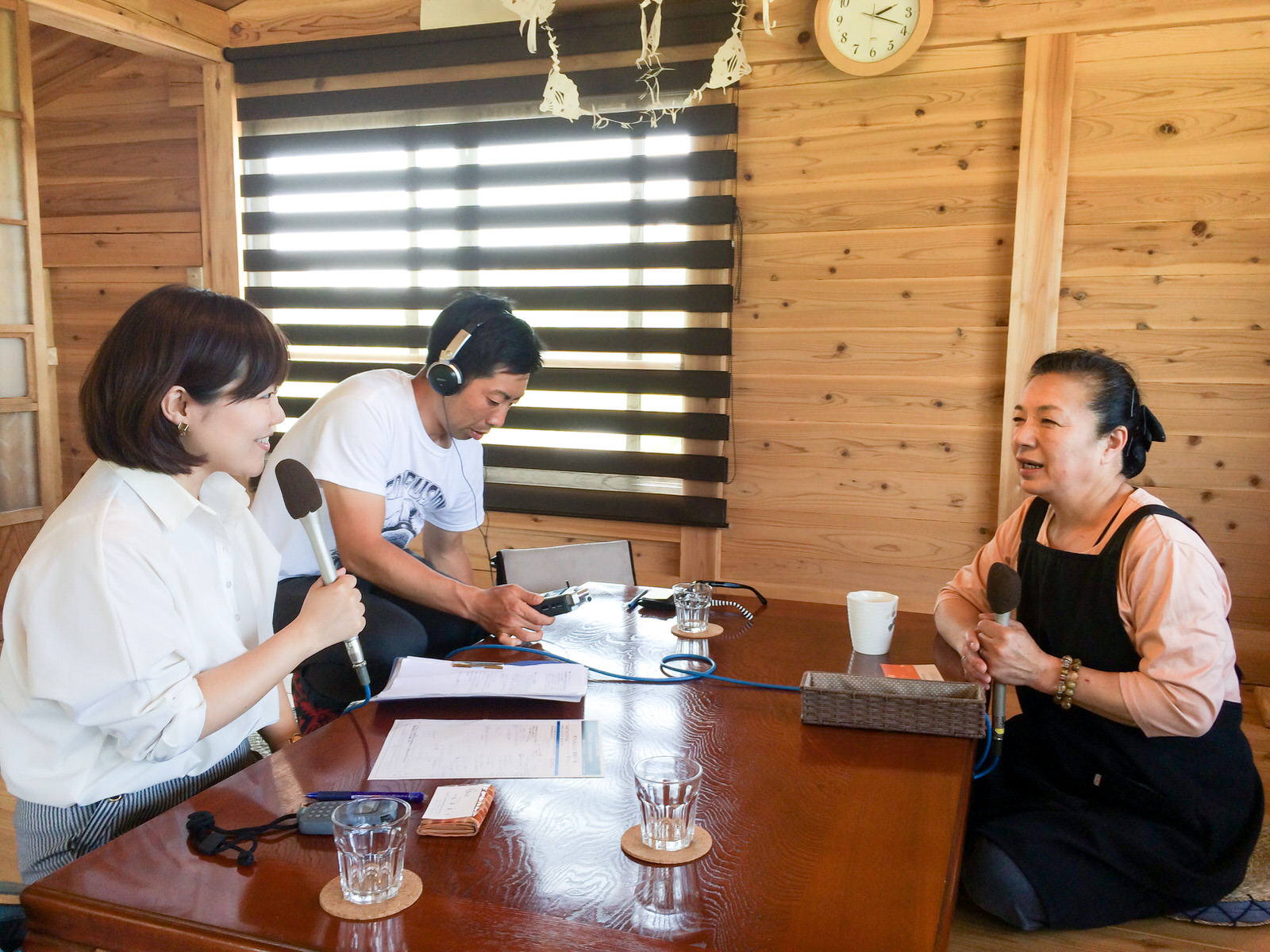 ヘッドホンをした男性の横でマイクを持って座っているリポーターの女性が、テーブルをはさんでマイクを持って座った成澤さんにインタビューをしている様子の写真