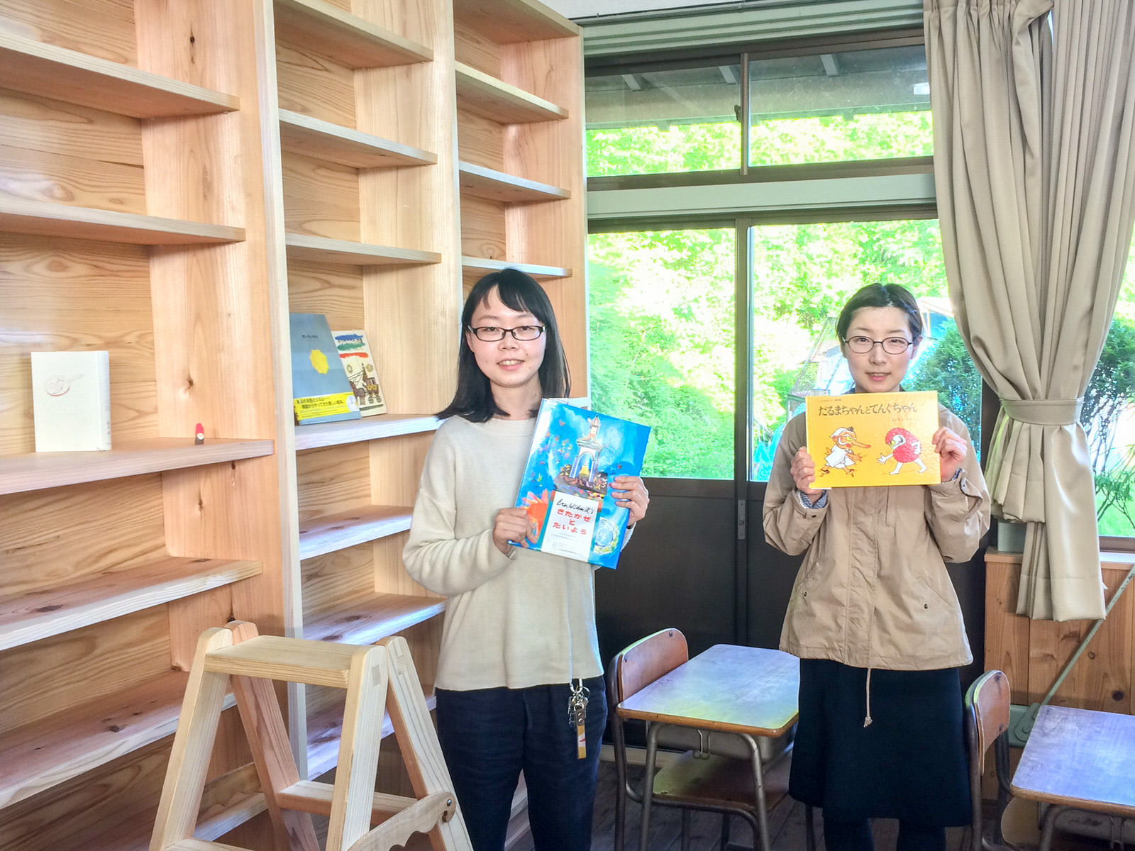 机といすが並ぶ教室の左側に大きな本棚があり、それぞれ本を持った二人の女性が並んで立っている写真