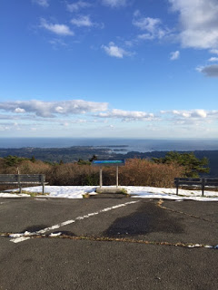 アスファルトの展望スペースにうっすらと雪が残る中、正面に設置された青い案内板と左右にベンチがあり、その向こうに森と海が遠くまで見渡せる景色の写真