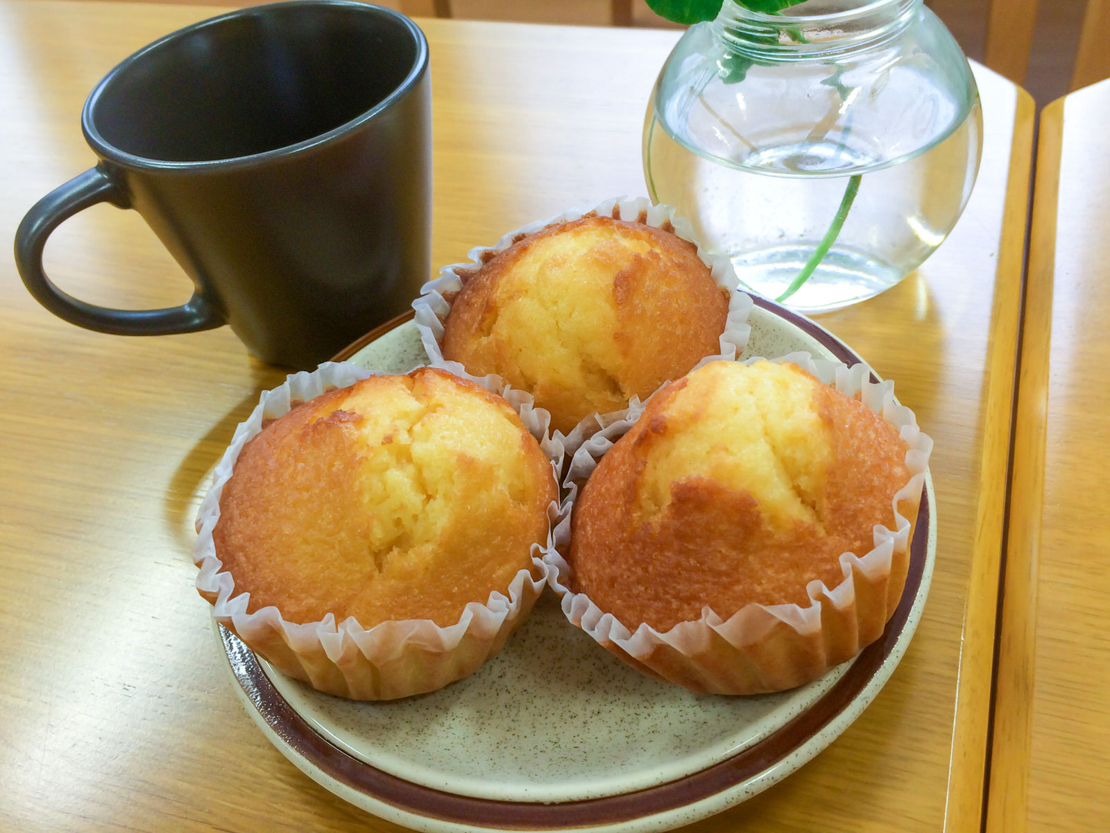 丸い皿に、ふっくらとしたきつね色のマドレーヌが3つ盛り付けられ、マグカップと花瓶が添えられている写真