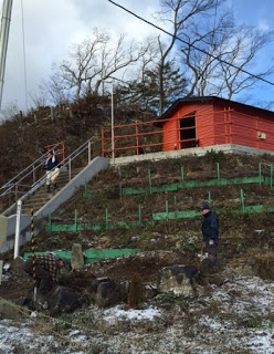 雪が残る寒そうな斜面で、赤い建物へと続く階段の横に立つ人物と、緑色のネットやフェンスが張られた地面で掃除をしているもう1人の人物の様子を写した写真
