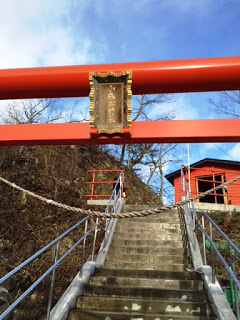 青空の下、石段の上に朱色の鳥居が立ち、中央の額には「八幡神社」と書かれており、鳥居をくぐった先の階段の上には赤い建物が建っている様子の写真
