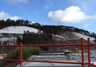 空には雲が浮かびつつ青空も見え、その先には雪に覆われた斜面と森林が広がる丘陵地帯があり、赤い手すりの向こうに雪が積もった小高い山が見える風景の写真