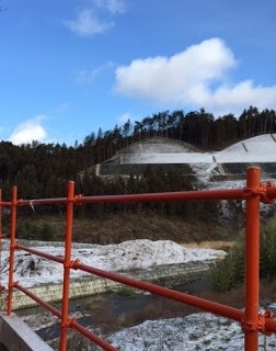 空には雲がありつつ青空も見え、背景には雪に覆われた丘と木々が広がり、赤い金属製の手すりの向こうには、雪に覆われた景色の中を八幡川が静かに流れている写真