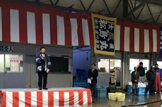 紅白幕で飾られたステージ上で、男性がマイクを使って話しており、周囲には話を聞いているような人々が立っていて、背景には大漁旗や箱・容器などが並んでいる、初競りの様子を写した写真