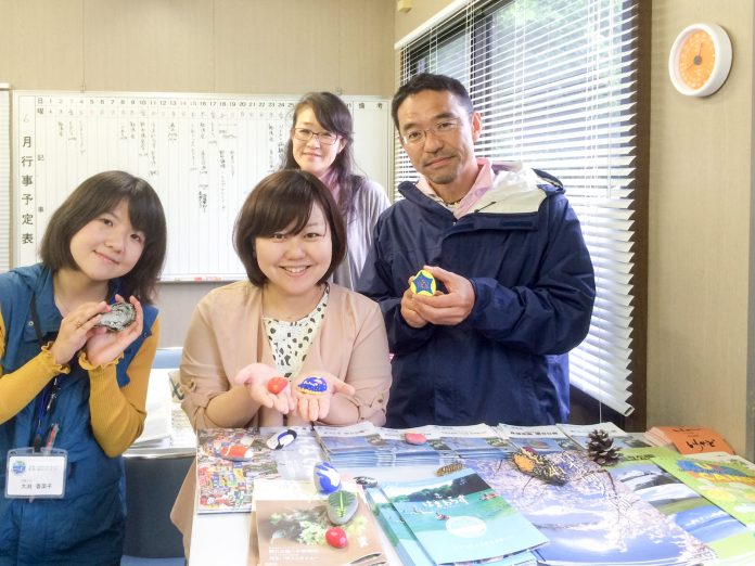 リーフレットなどがたくさん並んだカウンターの後ろに立った女性2人と男性が、カラフルにペイントした石を手に持って、後ろに立つ女性と一緒に写した写真
