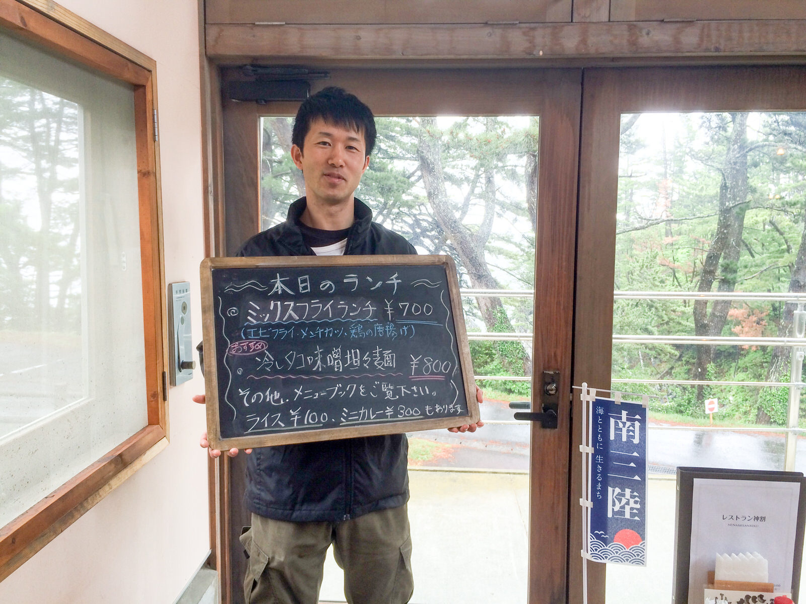 外に緑の木々が見える木の枠のガラス窓の前で、及川さんが本日のランチのメニューが書かれた黒板を胸の前で持って立っている写真