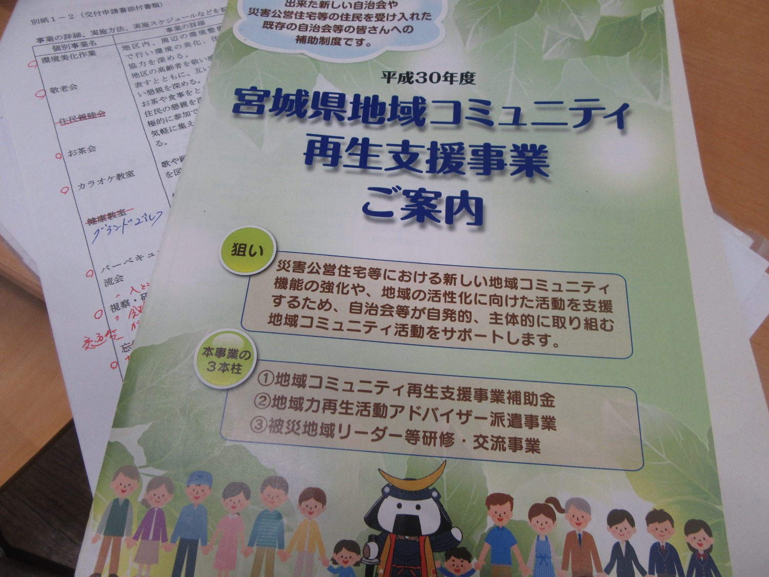 「平成30年度 宮城県地域コミュニティ再生支援事業ご案内」と書かれたパンフレットが机の上に置かれ、災害公営住宅での地域活動支援について説明されており、下には手書きの赤字が書き込まれた活動計画表のような資料が見える写真