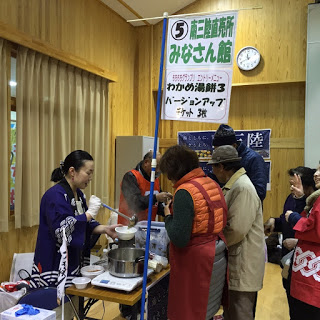 南三陸直売所みなさん館ののぼりが立ったテーブルで、濃い紫色の法被を着た女性が鍋から温かいわかめ湯餅をお椀によそい、テーブル前には商品を求める人々が集まっている様子の写真