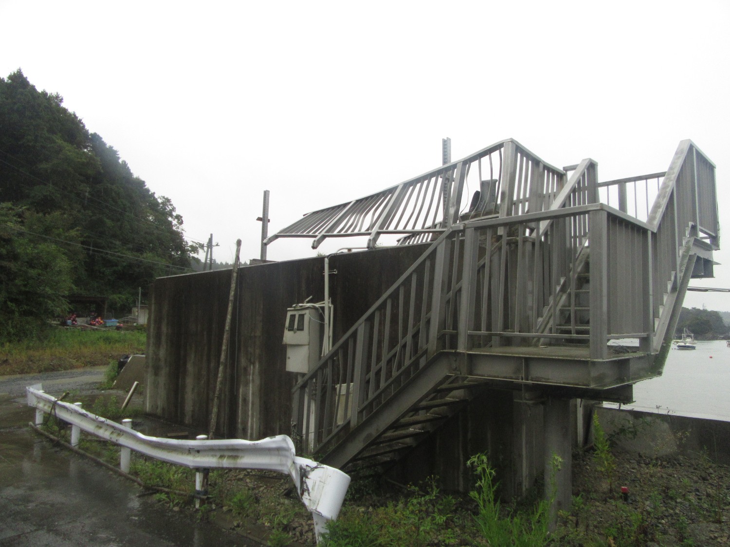 川沿いの道路脇に設置された鉄製の階段が、上部の手すりや屋根部分を激しく損傷したまま放置されており、傍らには曲がったガードレールと濡れたアスファルトが見える写真