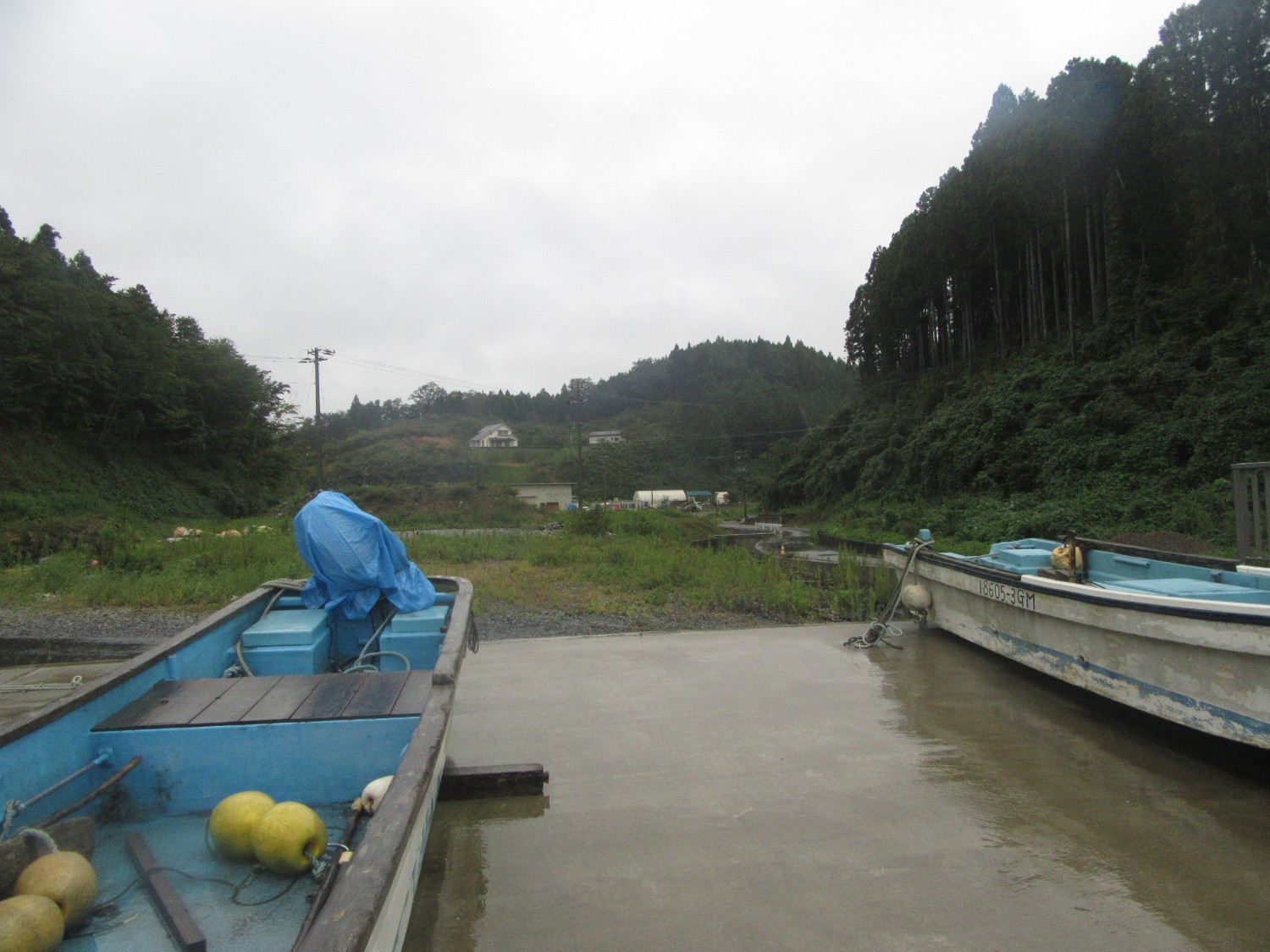 曇り空のもと、森に囲まれた静かな川べりに小型の漁船が数隻停められ、船の中には黄色い浮きや青いシートに覆われた機材が見える写真