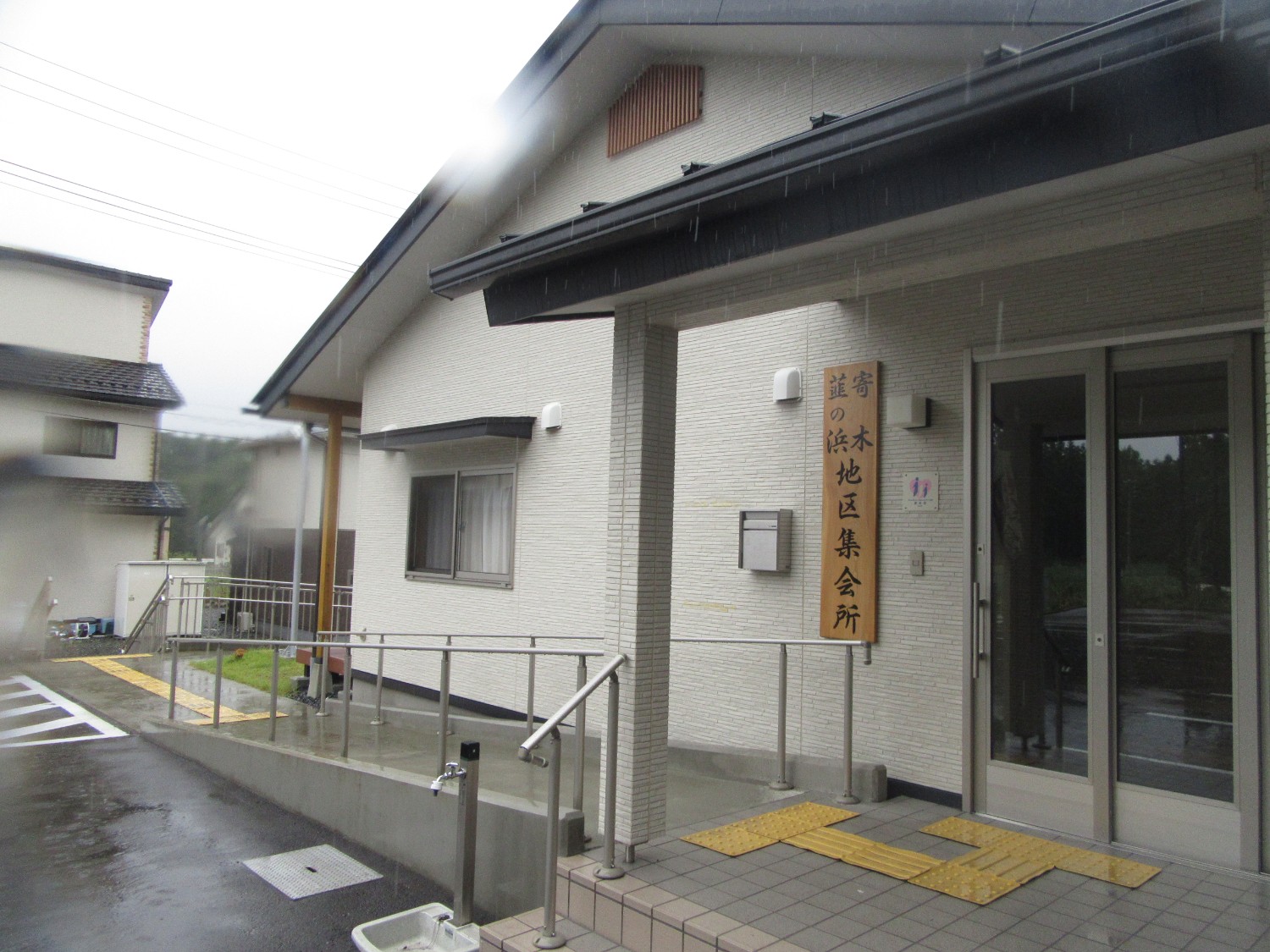 雨が降る中、白い外壁の平屋建ての建物の入口に「寄木・韮の浜地区集会所」と書かれた木の看板が掛けられ、バリアフリー対応のスロープと手すりが備えられている写真