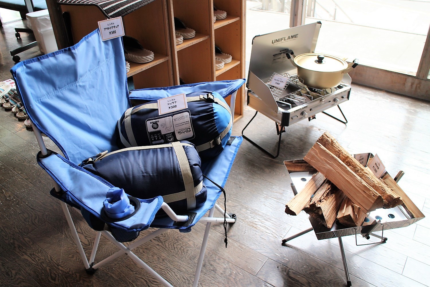 室内に置かれた青いアウトドアチェアの上にシュラフが2つ置かれ、隣にはキャンプ用ガスコンロと薪が準備されているレンタル用品コーナーの写真