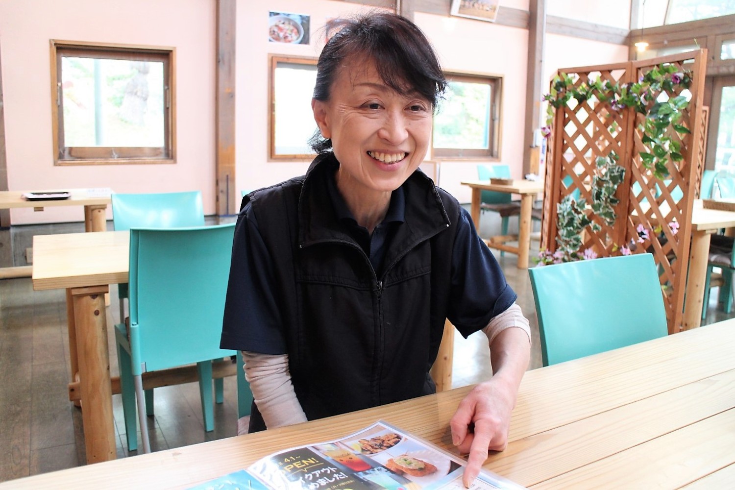 緑の椅子に木製テーブルが設置された屋内の明るいレストラン内で、笑顔でメニューを指差している佐藤さんの写真