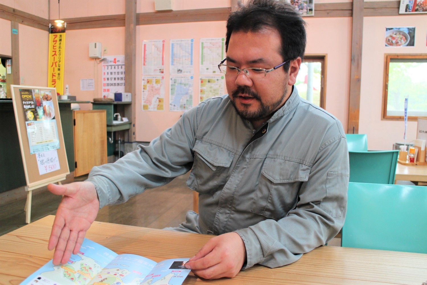 背景にはカレンダーや写真などが壁に掲示され、立て看板にメニュー表が貼られたレストラン内で、作業着姿の遠藤さんが木製テーブルに広げた地図を指差しながら説明している写真