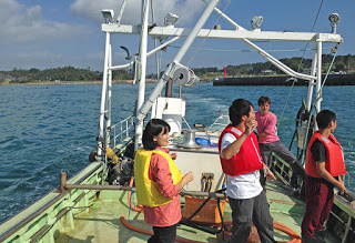 漁船の上にライフジャケットを着用した男女3人と、ツアーガイドの男性が乗っている写真