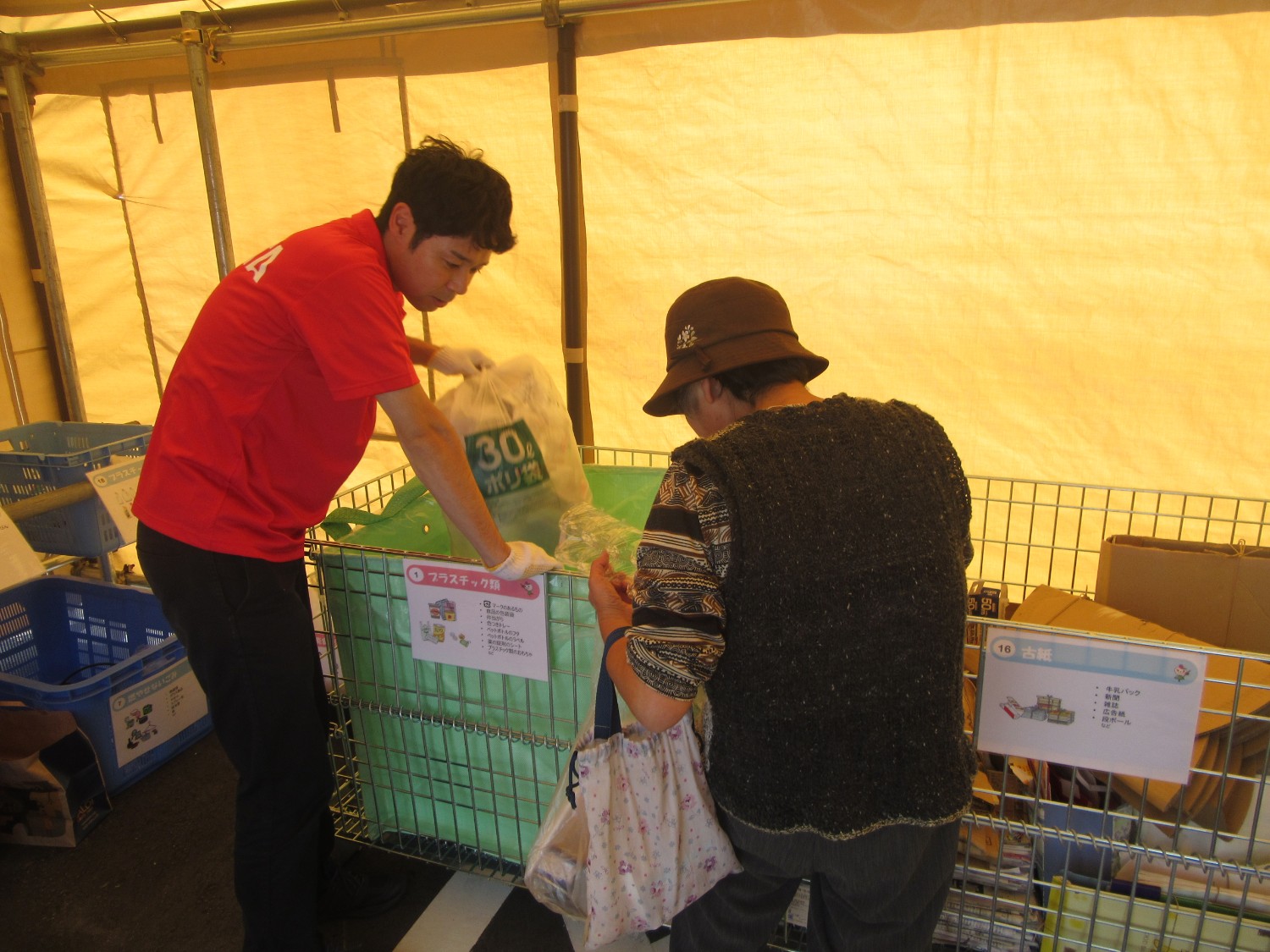 テント内で赤いシャツを着たスタッフが「プラスチック類」と書かれた回収カゴにポリ袋を投入しており、隣では帽子をかぶった高齢の女性が「古紙」回収カゴの前でごみを仕分けしている様子の写真