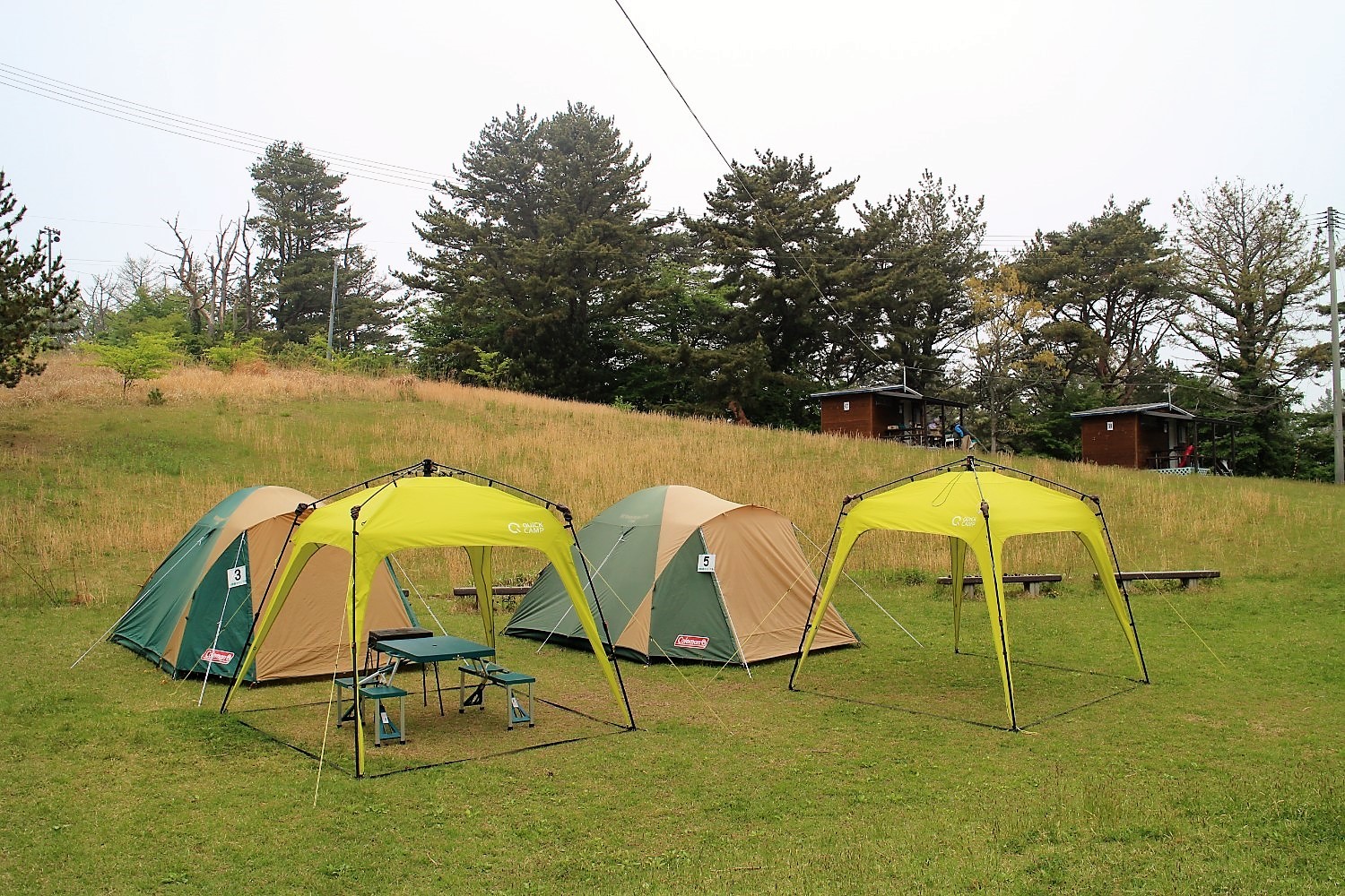 緩やかな斜面の草原に緑とベージュのテントが二つ並び、その前に黄緑色の簡易屋根とピクニックテーブルが設置され、後方には木造のキャビンが見えるキャンプサイトの写真