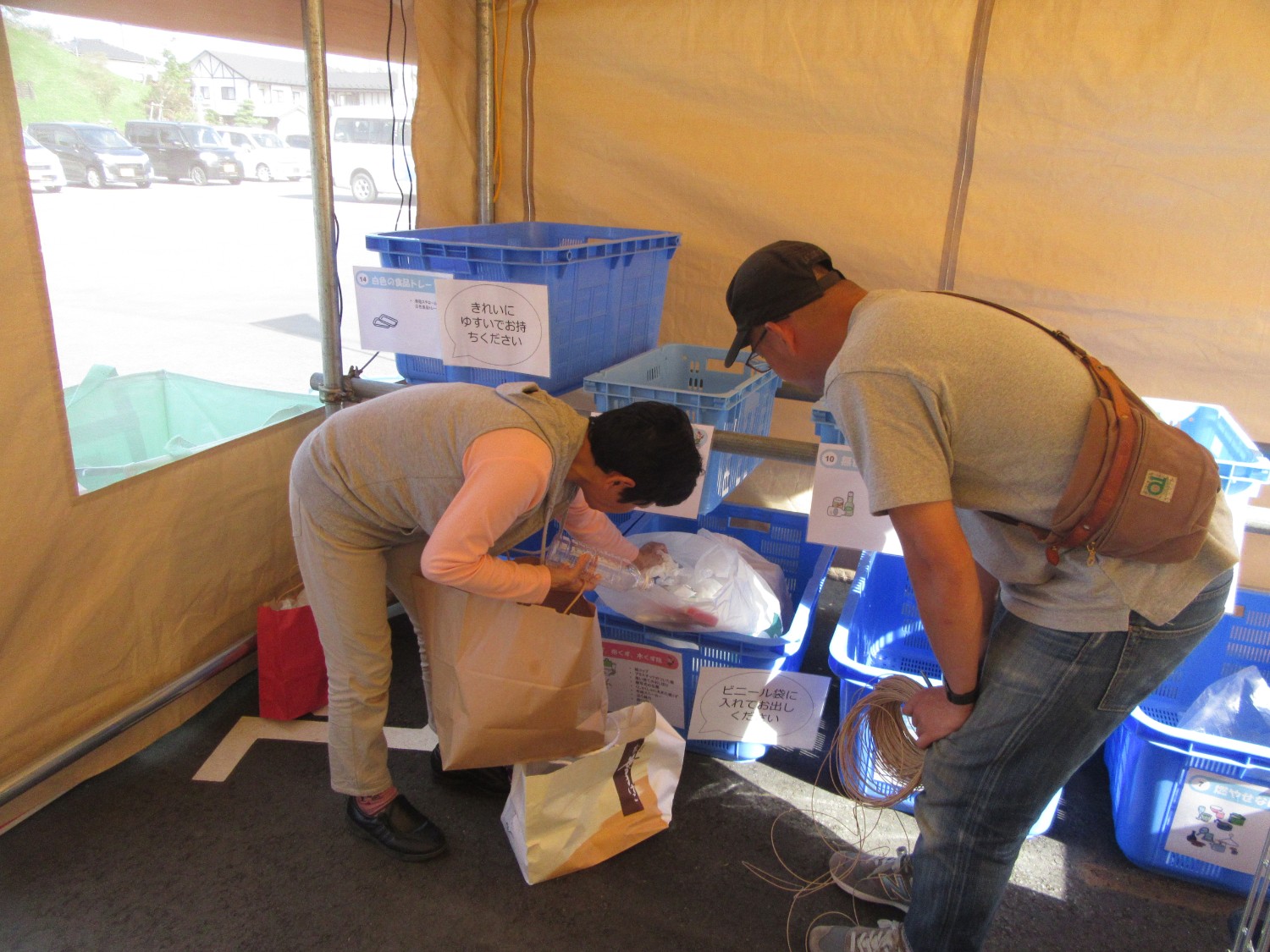 テントの中で、青い回収ボックスの前に立つ2人の人物が紙袋からごみを取り出して分別作業をしており、ボックスには「きれいにゆすいでお持ちください」などの分別指示が書かれた紙が貼られている様子の写真