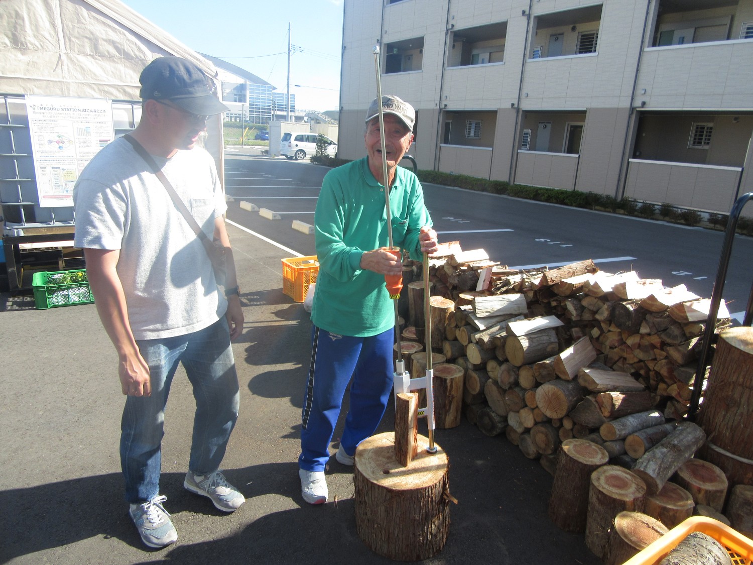 アスファルトの上で、薪割り器を使って丸太を割ろうとしている高齢の男性と、その様子を見守る帽子をかぶった男性が写っており、背後にはきれいに積まれた大量の薪と集合住宅が見える写真
