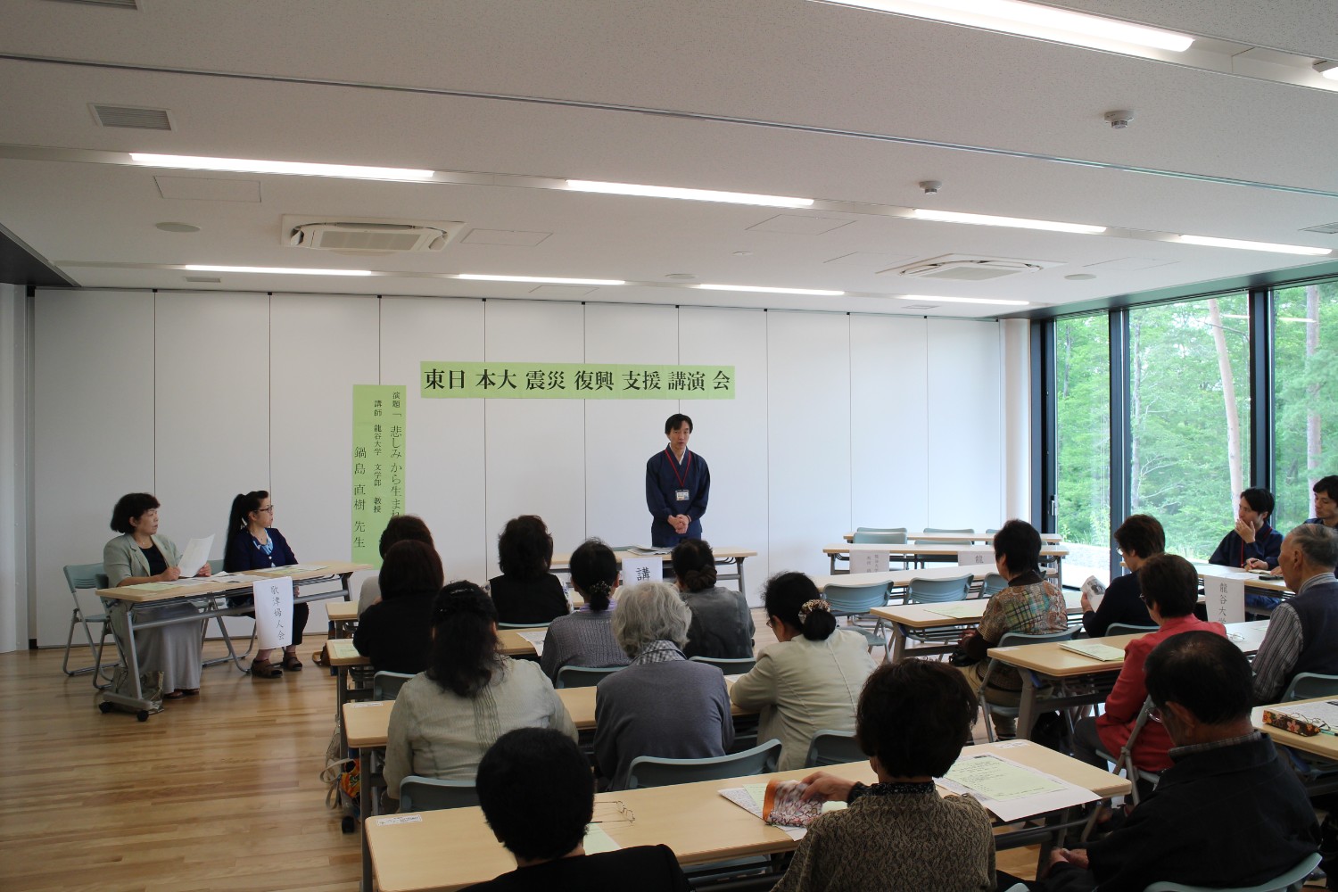 室内前方に講師が立ち参加者に向かって話しかけており、全員が真剣に耳を傾けている東日本大震災復興支援講演会の様子の写真