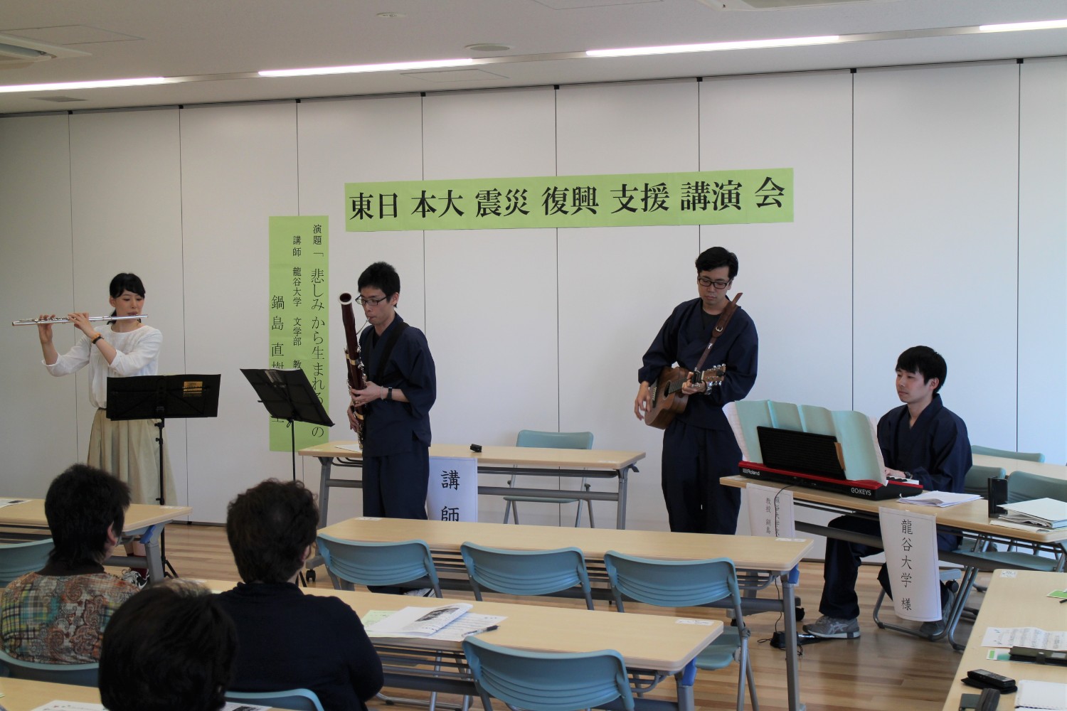 講演会が行われている室内で、フルート・ファゴット・ギター・キーボードの四人編成で演奏が行われており、観客が静かに聴いている音楽演奏の写真