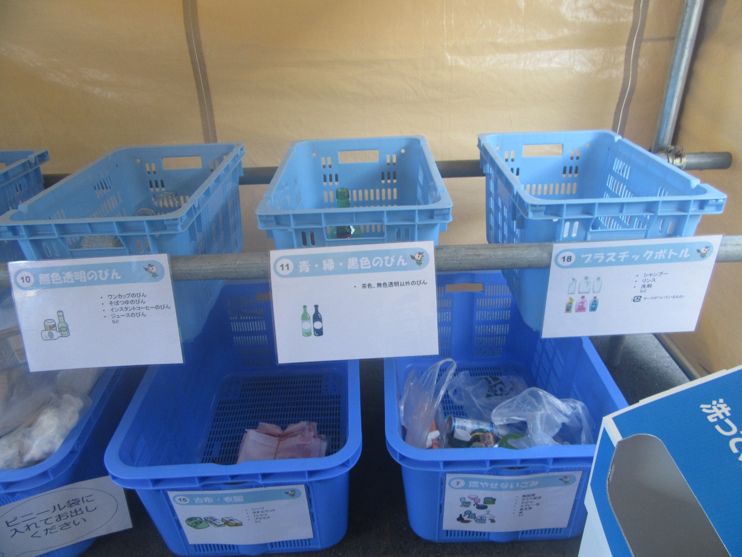 テント内の一角に青いプラスチック製の回収ボックスが並べられ、それぞれ「無色透明のびん」「青・緑・黒色のびん」「プラスチックボトル」などの分類ラベルとイラスト付きの案内が表示されており、瓶や容器が分別されている様子の写真