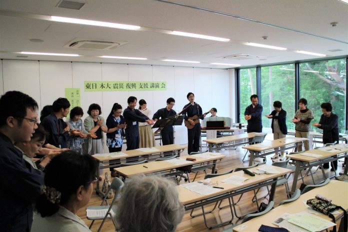 「東日本大震災 復興支援講演会」と書かれた張り紙がされている室内で、講師と参加者が輪になって広がり、手を前に出しながら音楽に合わせて合唱している様子の写真