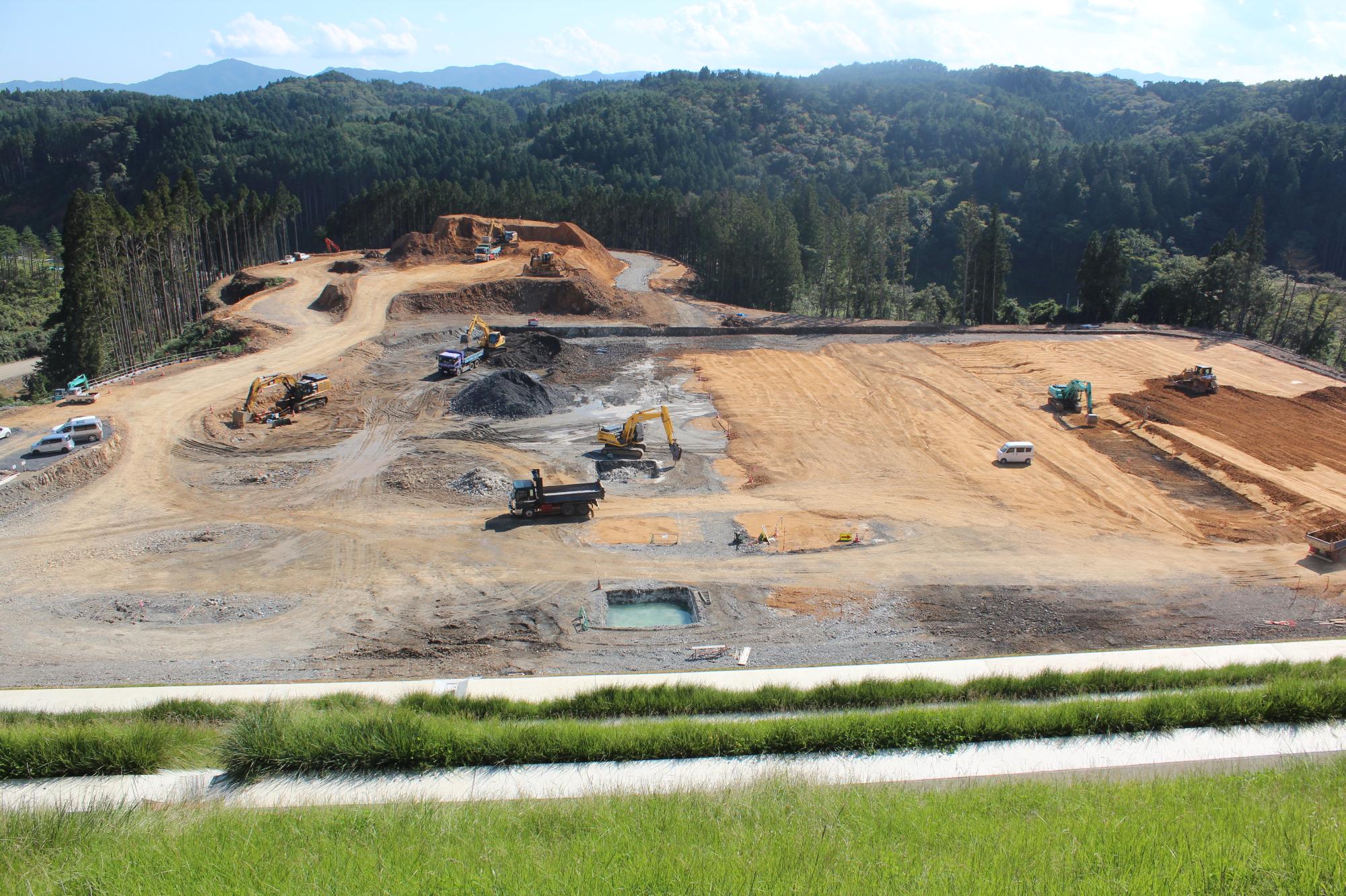 山の斜面に沿って工事が展開され、土砂運搬トラックやショベルカーが複数台稼働している造成地の写真