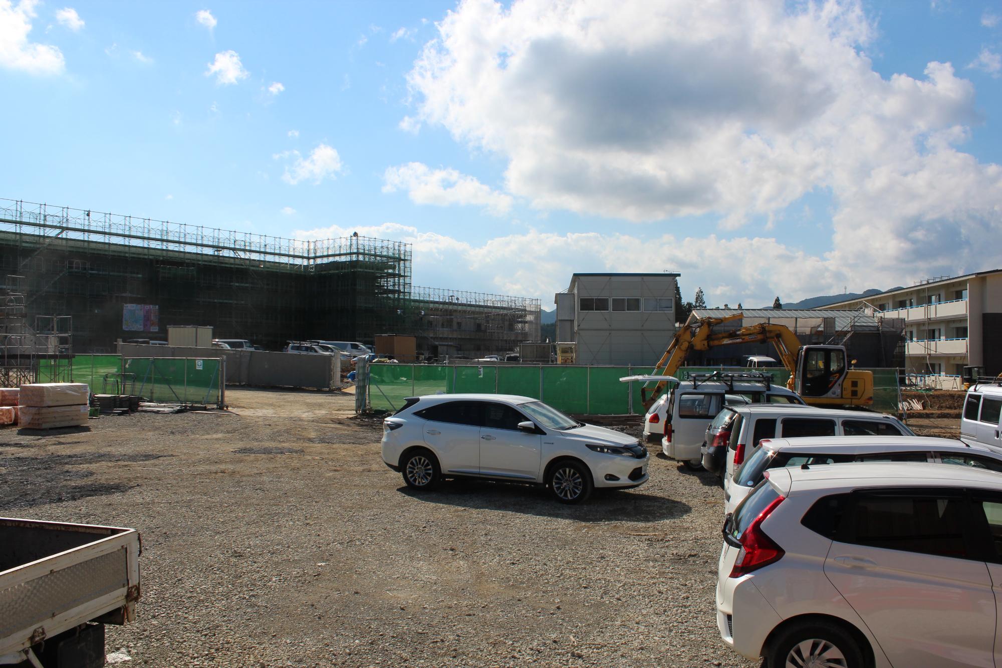 駐車された複数の車やショベルカーの手前に、仮設建物と足場の組まれた建設中の施設がある晴天の建設現場の写真
