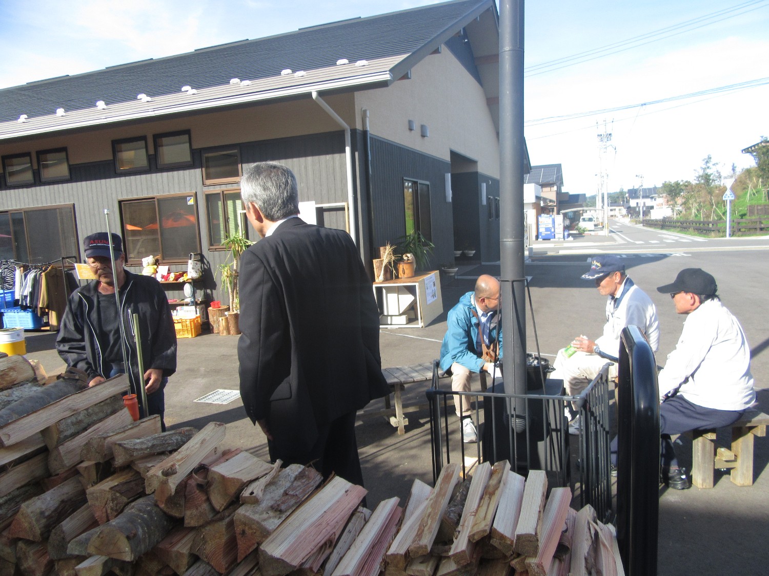 晴れた日の屋外で、薪ストーブの周りにベンチを囲んで座る高齢の男性たちや、薪を前に立って会話する人々が写っており、背景には黒とベージュの建物やリサイクル品らしき品物が並ぶスペースがある写真