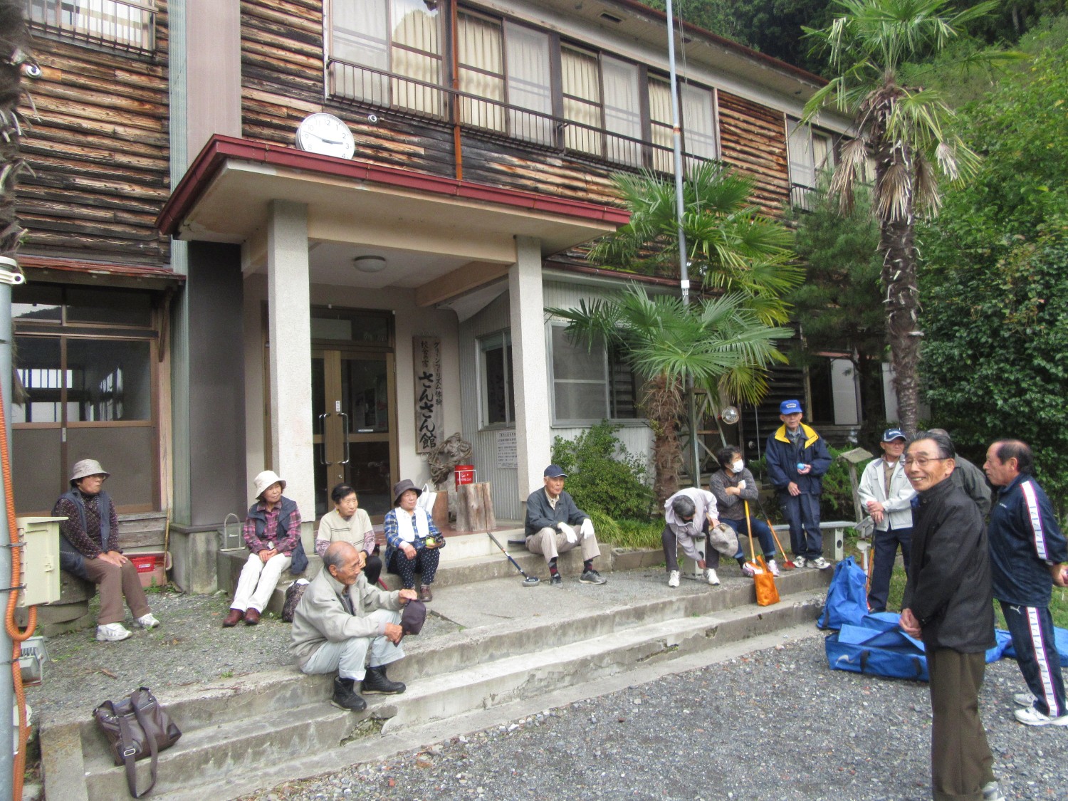 年配の男女が建物の前に集まり、くつろいでいる様子の写真