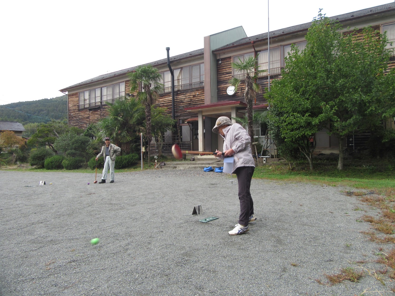 高齢の男女が校庭のような広い砂地でグラウンドゴルフを楽しんでいる写真