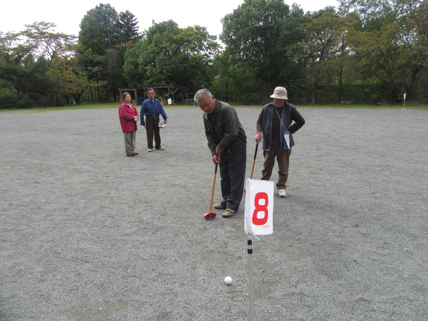 屋外の広い砂利のグラウンドで参加者が見守る中、年配の男性が8番の旗が付いたポールに向かってボールを打った写真