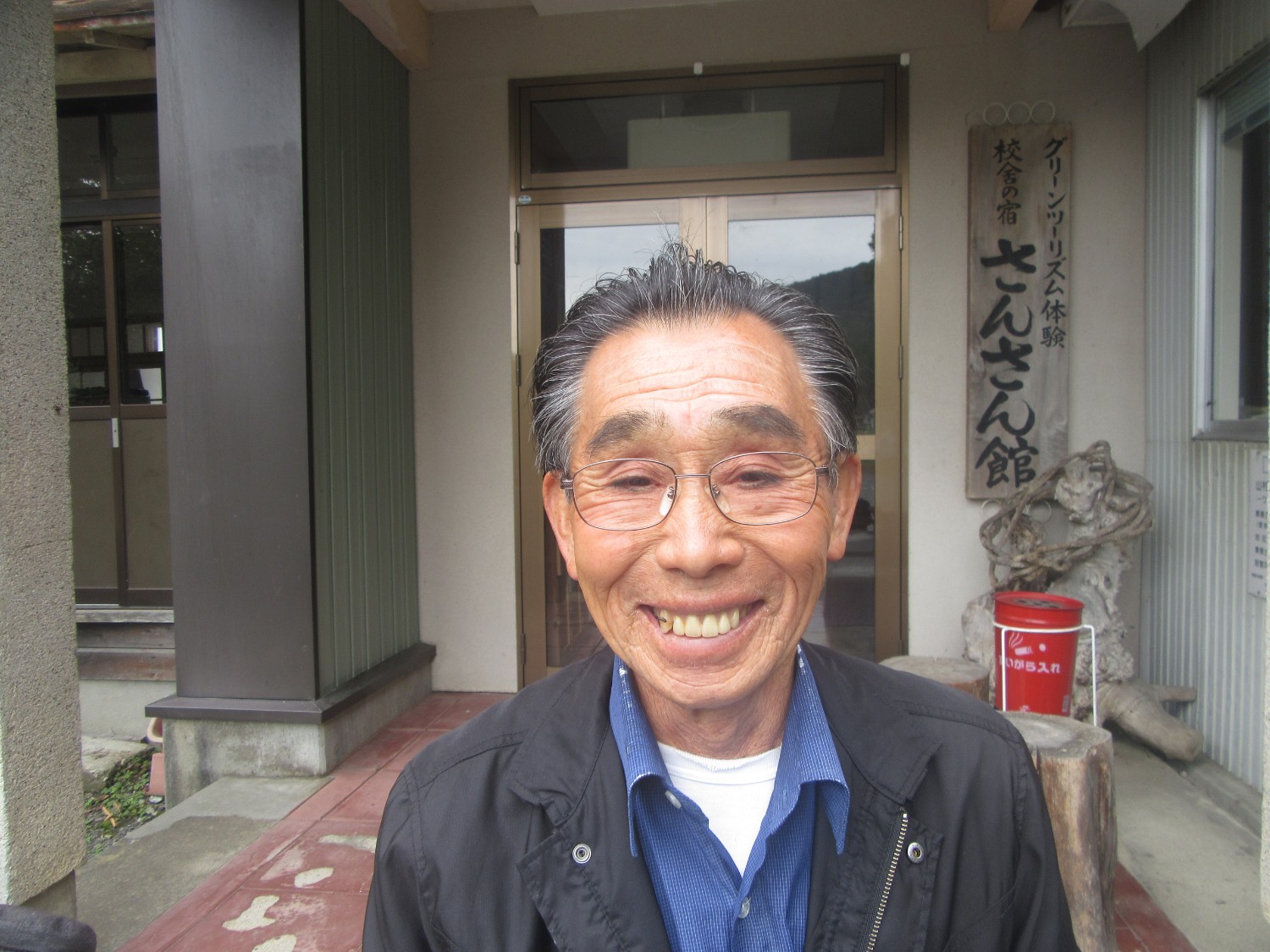 笑顔でカメラを見つめる眼鏡をかけた阿部忠雄さんが、さんさん館建物の入り口前に立っている写真